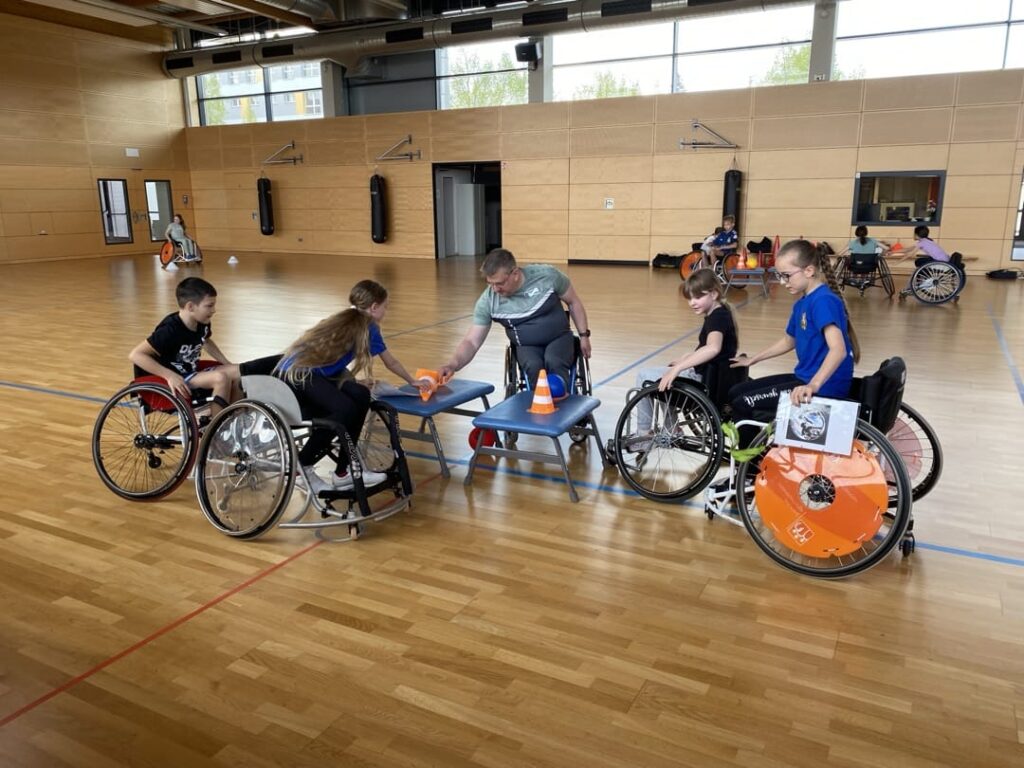 Kinder und ein Mann sitzen im Rollstuhl in einer Sporthalle. Der Mann zeigt einem Mädchen etwas.