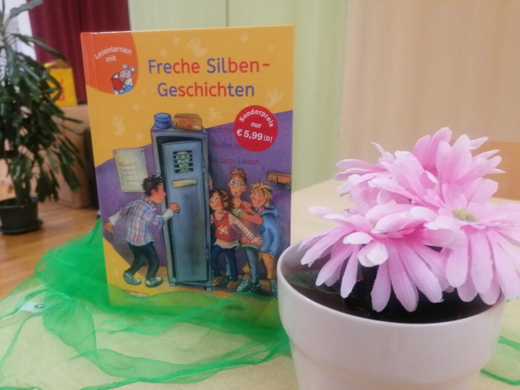 Eine weiße Vase mit rosa Blume auf einem Tisch. Daneben steht das Buch "Freche Silben Geschichten".