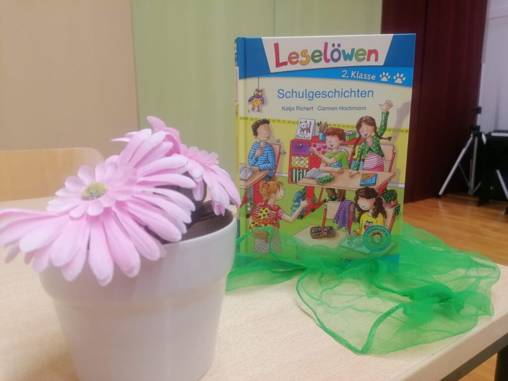 Eine weiße Vase mit rosa Blume auf dem Tisch. Dahinter steht ein Buch das Schulgeschichten heißt.