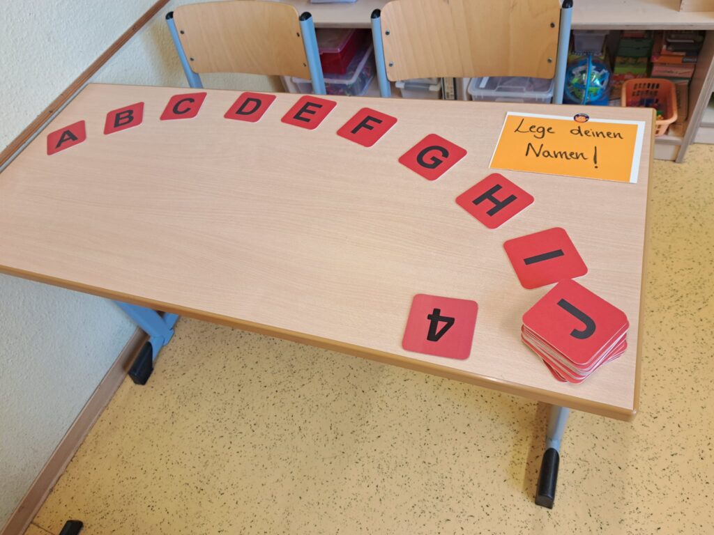 Tisch auf dem Karten liegen mit einzelnen Buchstaben darauf. Oben ein Schild mit der Aufforderung: Lege deinen Name!