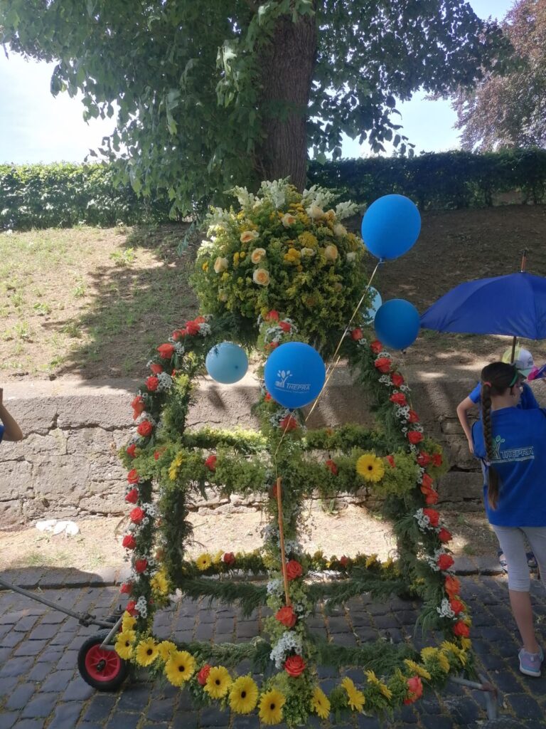 Ein kleiner Umzugswagen der aus Blumen und Pflanzen besteht, an diesem hängen blaue Luftballons.