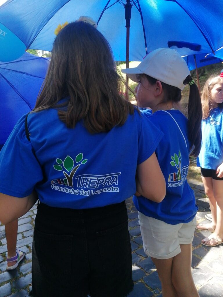 Zwei Mädchen mit blauen T-Shirt von der THEPRA Grundschule und blauen Regenschirm.