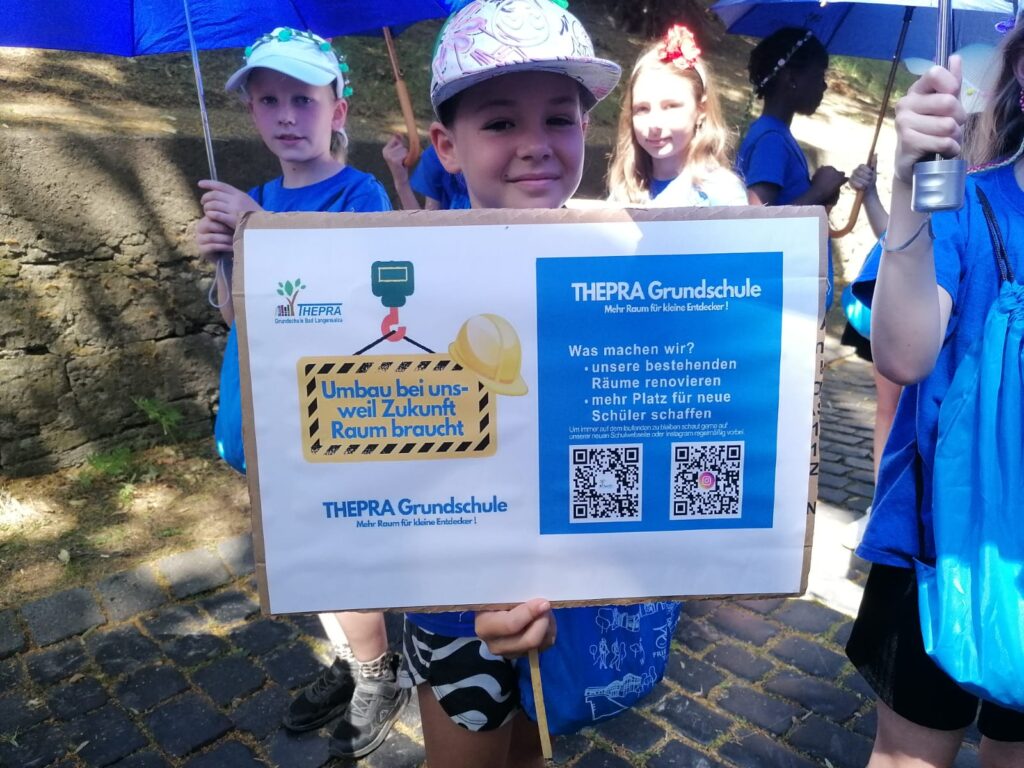 Junge mit Mütze zeigt beim Umzug ein Schild hoch. Dieses sagt, dass die THEPRA Grundschule umbaut um mehr Platz für neue Schüler zu haben.