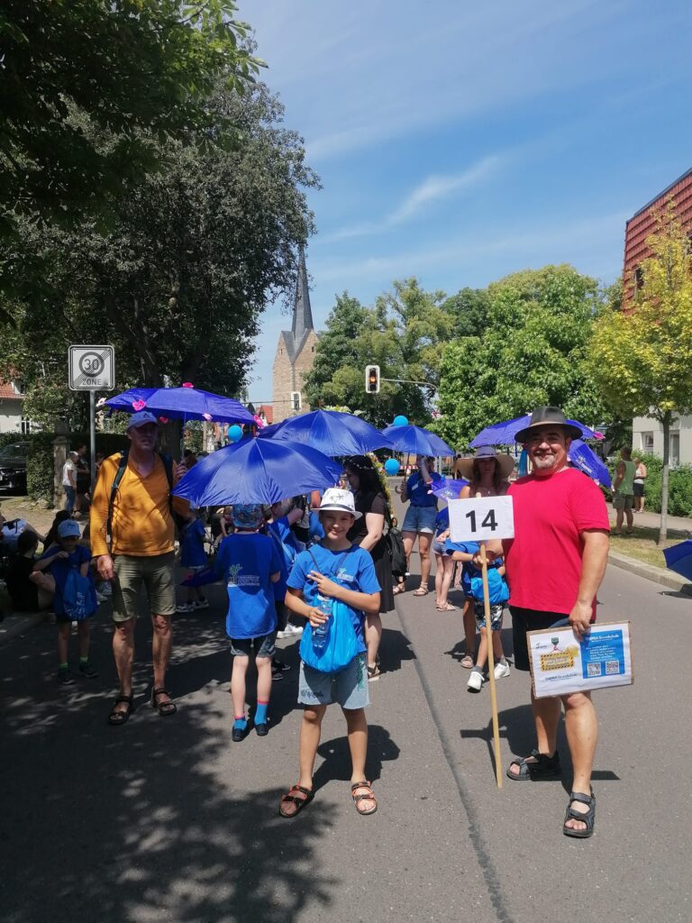 Junge mit blauen THEPRA T-Shirt und Regenschirm zeigt glücklich Peace. Mann mit rotem T-Shirt steht daneben mit einem Schild das 14 zeigt.